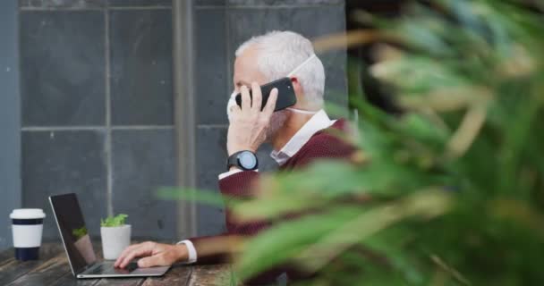 中年男子白种人坐在咖啡台上的桌子旁 头戴口罩 头戴眼镜 头戴眼镜 手持智能手机和笔记本电脑 动作缓慢 — 图库视频影像