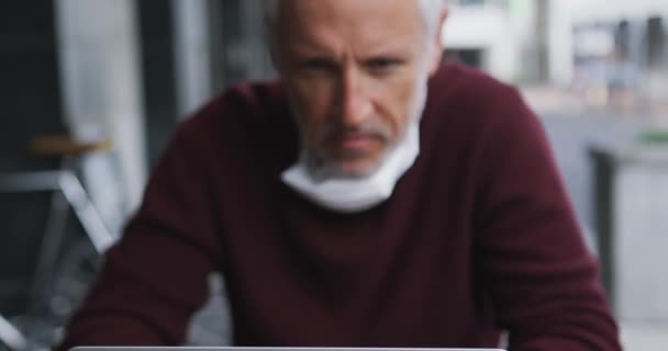 コーヒーテラスのテーブルに座っている中年の白人男性 コロナウイルスに対して顔マスクを身に着けています Covid 彼のラップトップコンピュータを使用して スローモーションで — ストック動画