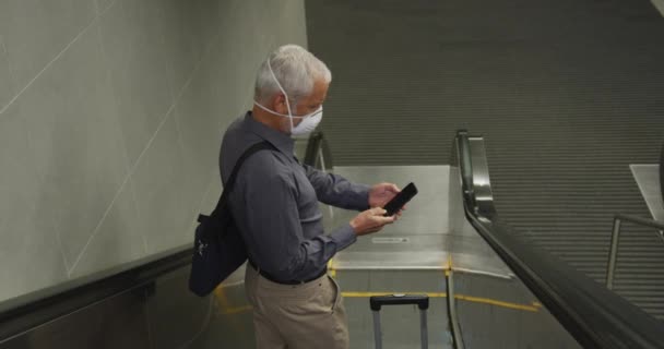 地下鉄の駅でエスカレーターを使用して 彼のスマートフォンを使用して スローモーションで コロナウイルスに対して顔マスクを身に着けている中年の男性 Covid — ストック動画