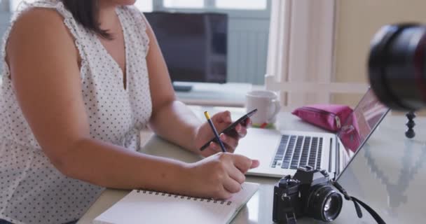 白人女性のVloggerは自宅で時間を過ごし テーブルに座ってノートパソコンやスマートフォンを使ってブログのビデオを準備している 隔離された封鎖における社会的距離と自己隔離 — ストック動画