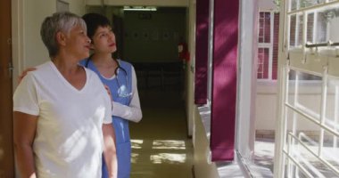Yaşlı melez bir kadın, güneşli bir günde pencereden dışarı bakıp konuşan beyaz bir kadın doktorla birlikte, Coronavirus covid19 salgını sırasında koridorda duruyor. Ağır çekimde