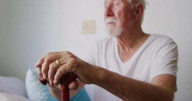 Elinde baston tutan yaşlı bir beyaz adamın Coronavirus covid19 salgını sırasında bir huzurevindeki odasında izole edilmiş ve sosyal mesafeli bir şekilde oturmasını. Ağır çekimde