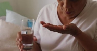 Yaşlı melez bir kadın, bir huzurevinde yatağında oturmuş, Coronavirus covid19 salgını sırasında bir bardak suyla ilacını alan beyaz bir kadınla konuşuyor..