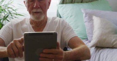Beyaz sakallı, gülümseyen ve tablet bilgisayar kullanan, Coronavirus Covid19 salgını sırasında bir huzurevindeki yatak odasında sosyal mesafeli ve izole edilmiş yaşlı bir beyaz adamın fotoğrafını çek.