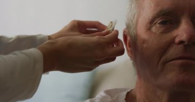 Bir huzurevindeki yaşlı beyaz bir adamın Coronavirus covid19 salgını sırasında beyaz bir kadın hemşire tarafından oturtulması ve işitme cihazıyla donatılması..