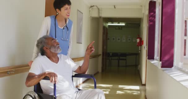 老年男女混血男子坐在走廊的轮椅上 与一位白人女医生坐在养老院里 在一个阳光明媚的日子里 在考罗内韦尔斯病流行的时候 他们一起笑着 说话着 凝视着窗外 — 图库视频影像