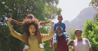 Çoklu nesil melez bir aile bahçede eğleniyor, karışık ırktan bir kız annesinin omuzlarında oturuyor ve melez bir çocuk dedelerinin omuzlarında oturuyor, el ele tutuşuyor ve gülümsüyor, ağır çekimde.