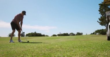 Güneşli bir günde golf sahasında duran, şapkalı ve şortlu, golf sopası sallayan ve yavaş çekimde topa vuran beyaz bir erkek golfçünün arka görüntüsü.