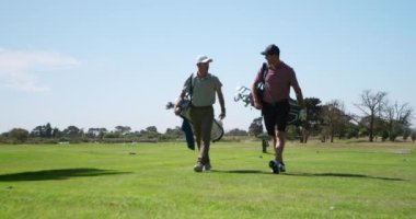 Güneşli bir günde golf sahasında yürüyen iki beyaz erkek golfçü şapkalar ve golf kıyafetleri giyip golf sopaları taşıyor ve ağır çekimde gülümsüyor ve konuşuyorlar.