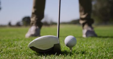 Alçak açı golf topunun bir golf sopasına yakın duruşu ve bir oyuncunun ayakları golf sopasını konumlandırıp geri sallaması güneşli bir günde golf sahasında başlama vuruşuna hazırlanması, ön plana odaklanması, yavaş çekim.