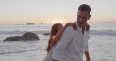 Side view of a happy young adult Caucasian couple relaxing by the sea at sunset, the woman jumping on the back of the man for a romantic piggyback, slow motion