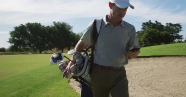 Kafkas erkek golfçü, güneşli bir günde golf sahasının yanından geçerken şapka ve golf kıyafetleri giyiyor, akıllı telefon kullanıyor ve ağır çekimde golf çantası taşıyor.