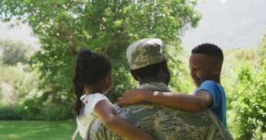 Karışık ırk adamı askeri üniforma giyiyor, eve dönüyor, bir bahçede duruyor, çocuklarını kucaklıyor ve kucaklıyorlar, çocuklar ellerinde küçük bayraklar sallıyor, ağır çekimde.