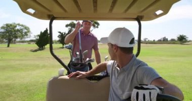 Güneşli bir günde golf sahasında iki beyaz erkek golfçü. Şapka ve golf kıyafetleri giyip oyun oynuyorlar. Golf çantasındaki bir sopayı değiştiriyorlar.