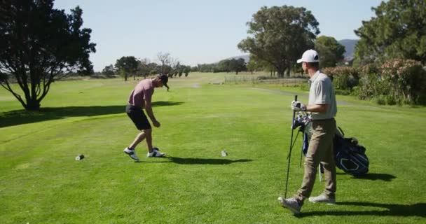 帽子とゴルフ服を着て晴れた日にゴルフコースに立っている2人の白人男性ゴルファー 一つは ティーの上にゴルフボールを置くために曲げオフに歯の前に 彼の対戦相手は ゴルフバッグを見て近くに立っている間 スローモーションで — ストック動画