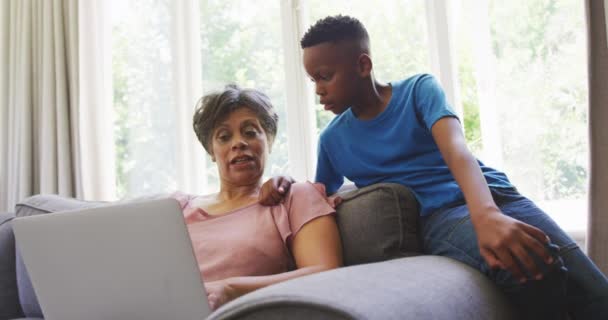 年龄较大的混血种族妇女享受在家里的时光 与孙子坐在沙发上 使用笔记本电脑 与外界保持距离 并在考拉病毒肆虐时自我隔离 动作缓慢 — 图库视频影像