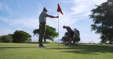 Güneşli bir günde golf sahasında iki beyaz erkek golfçü, bir oyuncu ayakta ve bayrak direğini tutarken, rakibi ağır çekimde topunu son deliğe atıyor.