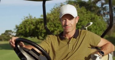 Güneşli bir günde golf sahasında golf şapkası ve golf kıyafeti giyen beyaz bir erkek golfçünün portresi. Golf arabasında oturuyor, kameraya gülümsüyor.