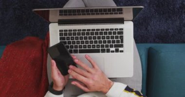 Kafkasyalı bir adamın evde vakit geçirmesini, sosyal mesafeyi ve kendini karantinaya almasını oturma odasındaki bir kanepede oturmasını akıllı telefon ve dizüstü bilgisayar kullanarak izlemesini.