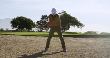 Güneşli bir günde golf sahasında şapkalı ve golf kıyafetli beyaz bir erkek golfçü, bir kum sığınağında duruyor, bir golf sopası sallıyor ve ağır çekimde topa vuruyor.