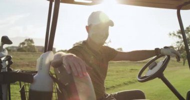 Güneşli bir günde golf sahasında golf şapkası ve golf kıyafetleri giymiş beyaz bir erkek golfçünün portresi golf arabasında oturur, kameraya döner ve gülümser.