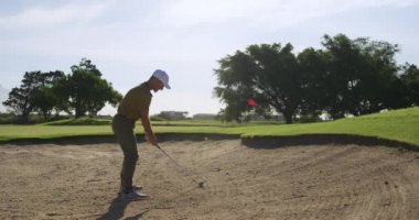 Güneşli bir günde golf sahasında şapkalı ve golf kıyafetli beyaz bir erkek golfçü, bir kum sığınağında duruyor, bir golf sopası sallıyor ve topa sığınaktan dışarı vuruyor, kırmızı bir bayrak direğiyle ağır çekimde deliğe doğru.