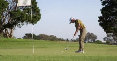 Güneşli bir günde golf sahasında beyaz bir erkek golfçü şapka ve golf kıyafetleri giyer, elinde golf sopasıyla topu son deliğe atar, ön planda bayrak direğiyle işaretler ve yavaş çekimde yumruğunu kaldırır.