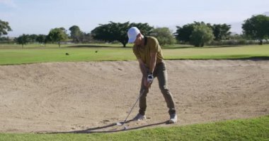 Güneşli bir günde golf sahasında şapkalı ve golf kıyafetli beyaz bir erkek golfçü, bir kum sığınağında duruyor, bir golf sopası sallıyor ve ağır çekimde topa vuruyor.