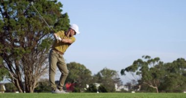 Güneşli bir günde, golf sahasında şapkalı ve golf kıyafetli, golf sopası sallayan ve topa yavaş çekimde vuran beyaz bir erkek golfçünün arka görüntüsü.