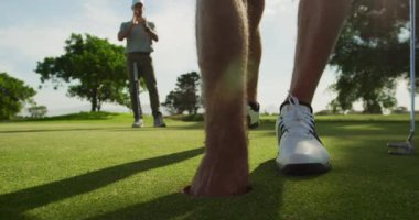 Güneşli bir günde golf sahasında şapkalı ve golf kıyafetli, elinde golf sopasıyla golf topunu delikten çıkaran, arka planda durup alkışlayan beyaz bir erkek golfçünün alçak açılı görüntüsü.