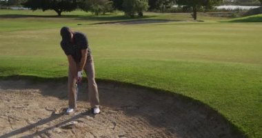 Güneşli bir günde golf sahasında şapkalı ve golf kıyafetli beyaz bir erkek golfçü, bir kum sığınağında duruyor, bir golf sopası sallıyor ve ağır çekimde topu sığınaktan deliğe doğru fırlatıyor.