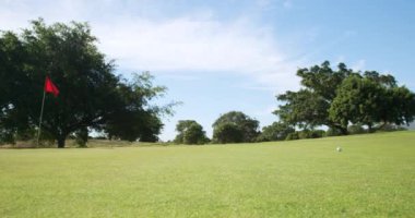 Kafkasyalı bir erkek golfçünün güneşli bir günde golf sahasına çömelmiş bir şapka ve golf kıyafetleri içinde elinde golf sopasıyla kırmızı bir bayrak direğiyle topu son deliğe sokmaya hazırlanırken yan görüntüsü.