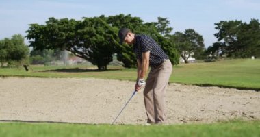 Güneşli bir günde golf sahasında şapkalı ve golf kıyafetli beyaz bir erkek golfçü, bir kum sığınağında duruyor, bir golf sopası sallıyor ve ağır çekimde topu sığınaktan deliğe doğru fırlatıyor.