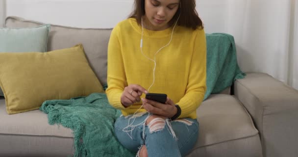 一名白人妇女呆在家里 与外界保持距离 自我隔离 被隔离 坐在客厅的沙发上 使用智能手机 戴耳机 — 图库视频影像