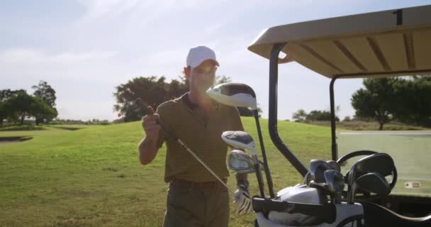 在阳光灿烂的日子里 白人男子高尔夫球手头戴高尔夫球帽 身穿高尔夫球衣 坐在高尔夫球车后面的高尔夫球袋里 准备慢吞吞地穿过高尔夫球场 — 图库视频影像