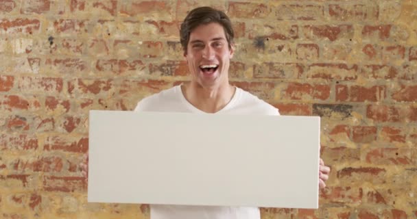一个白人男子的画像 手里拿着空白的白色帆布 看着相机 面带微笑 站在砖墙前 与外界保持距离 并在隔离中自我隔离 — 图库视频影像