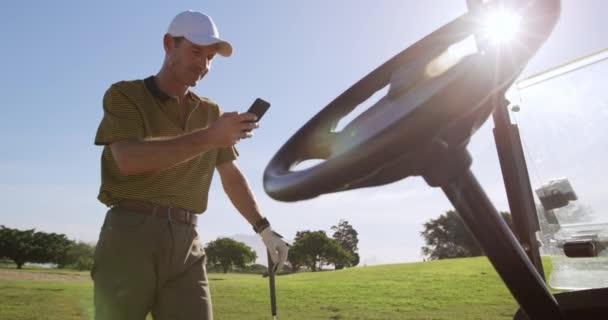 白人男性のゴルファーは 帽子とゴルフ服を着て晴れた日にゴルフコースを歩きます スマートフォンを使用して ゴルフクラブにもたれ 前景にゴルフバギーのステアリングホイール 日光によってバックライト スローモーションで — ストック動画
