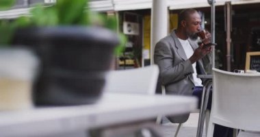 Afro-Amerikalı bir adam kafede vakit geçiriyor, kahve terasında oturuyor, bir fincan kahve içiyor ve akıllı telefonunu kullanıyor..