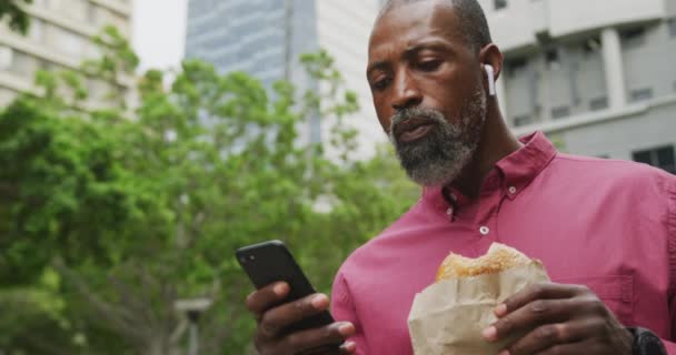 昼間はアフリカ系アメリカ人の男性が街中を歩き イヤフォンを身につけ スマートフォンを使いながらおやつを食べていました — ストック動画