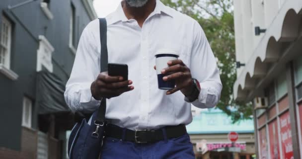 アフリカ系アメリカ人の男性が街を歩きながらテイクアウトコーヒーを飲みながらスマホを使っていました — ストック動画