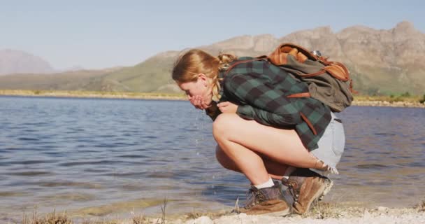 山への旅行に楽しい時間を過ごしている白人女性 湖畔に身をかがめて湖から水を飲み 晴れた日に景色を楽しむ スローモーションで — ストック動画