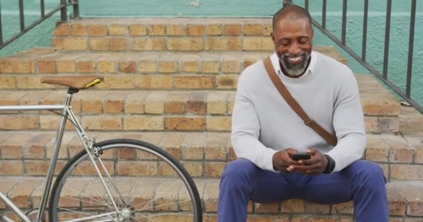 一日中アフリカ系アメリカ人の男性と街中を歩き 階段に座り 隣に自転車を置いてスマートフォンを使い — ストック動画