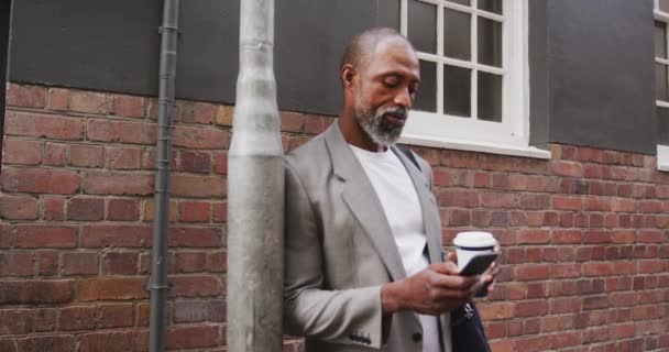 非裔美国人在城市街道上走来走去 站在街上 喝着外卖咖啡 用他的智能手机 — 图库视频影像