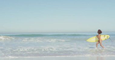Afrika kökenli Amerikalı bir kadının tropik bir plajda güneşin tadını çıkarırken, suda yürürken ve sarı sörf tahtasını ağır çekimde taşırken görüntüsü.