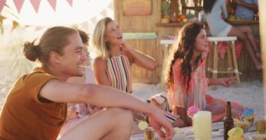 Gün batımında tropikal bir plajda oturup gitar çalarak ve yavaş çekimde gülerek eğlenen melez ırk arkadaşlarının ön görüntüsü.