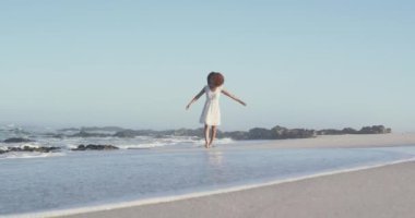 Afrika kökenli Amerikalı bir kadının, tropik bir plajda güneşin tadını çıkarması, yürümesi ve ellerini kaldırması ön planda.