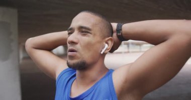 Melez bir ırk adamının yan görüntüsü. Şehir merkezinde çalışıyor, ara veriyor, ellerini başının arkasına koyup kulaklıkla, ağır çekimde müzik dinliyor.