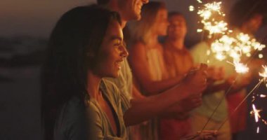 Gün batımında tropikal bir plajda eğlenen çok ırklı bir arkadaş grubunun yan görüntüsü. Yavaş çekimde maytaplarla gülümseyip oynuyorlar.