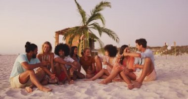 Gün batımında tropik bir plajda eğlenen, konuşan ve gülümseyen çok ırklı bir arkadaş grubunun ön görüntüsü.