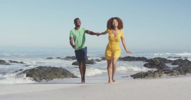 Afrika kökenli Amerikalı bir çiftin tropikal bir plajda güneşlenirken, yürürken, gülümserken ve el ele tutuşurken ağır çekimde